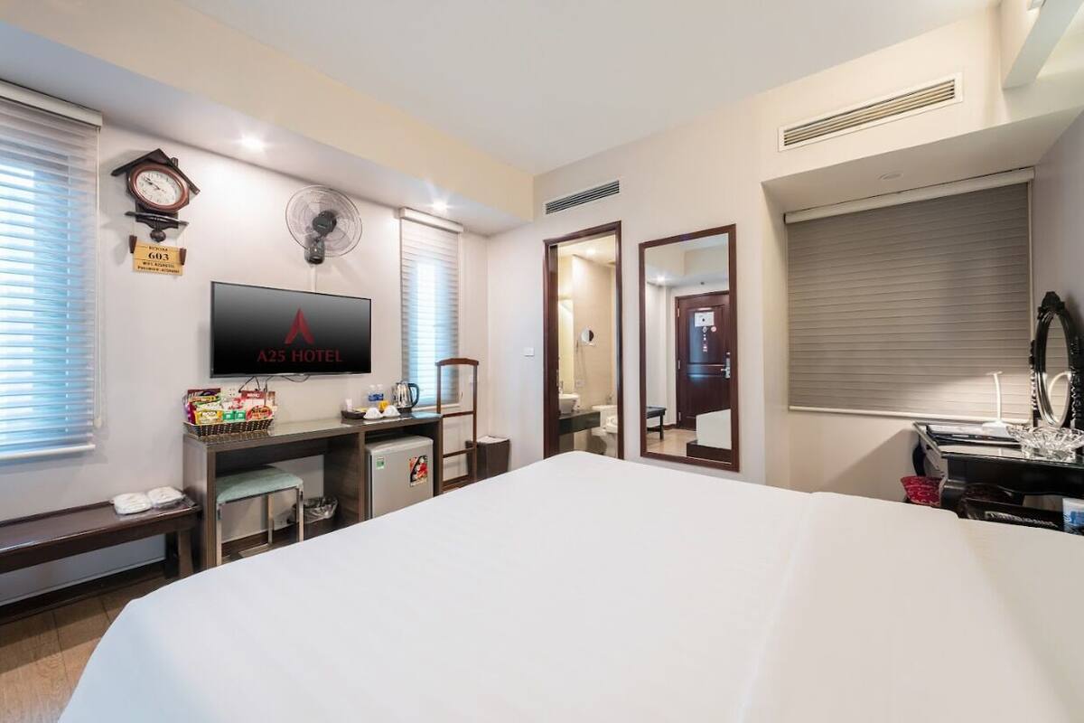 A hotel room with a large bed, a flat-screen TV, a desk with a computer, a small refrigerator, a clock, and a window with blinds.