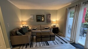 A living room with a sofa, a coffee table, and a patterned rug. There is a window with curtains and a view of the outdoors. -  Welcome to SSI...near Pier, Village and Beach! (St Simons)