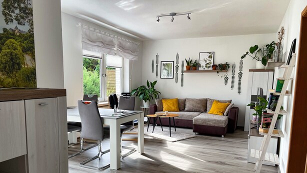 A modern living room with a dining area, wooden flooring, a sofa with yellow cushions, and a bookshelf with plants. - Ferienwohnung Im Kleinen Feld (Waldeck)