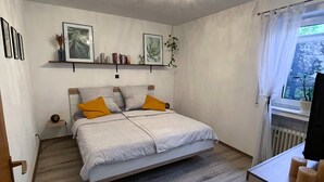 A bedroom with a bed, a bedside table, a shelf with books and a plant, and a window with curtains. - Ferienwohnung Im Kleinen Feld (Waldeck)
