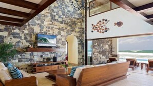 A spacious living area with a stone wall, a large flat-screen TV, wooden furniture, and a view of the ocean. - Fort Walker (Beachfront House) (Hacienda Iguana)