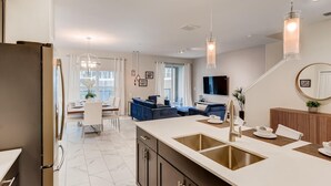 A modern kitchen with a central island, a dining area with a white table and chairs, and a living area with a blue sofa and a TV. - 5BR Villa Bliss: Private Pool - Sleeps 14 - 412 (Davenport)