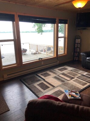 A room with a large window overlooking a dock and water, a wooden deck, a fireplace, a bookshelf, and a couch. - Barrett Lake, rent year round! Dock is out for the season as of 9.27.25 (Barrett)