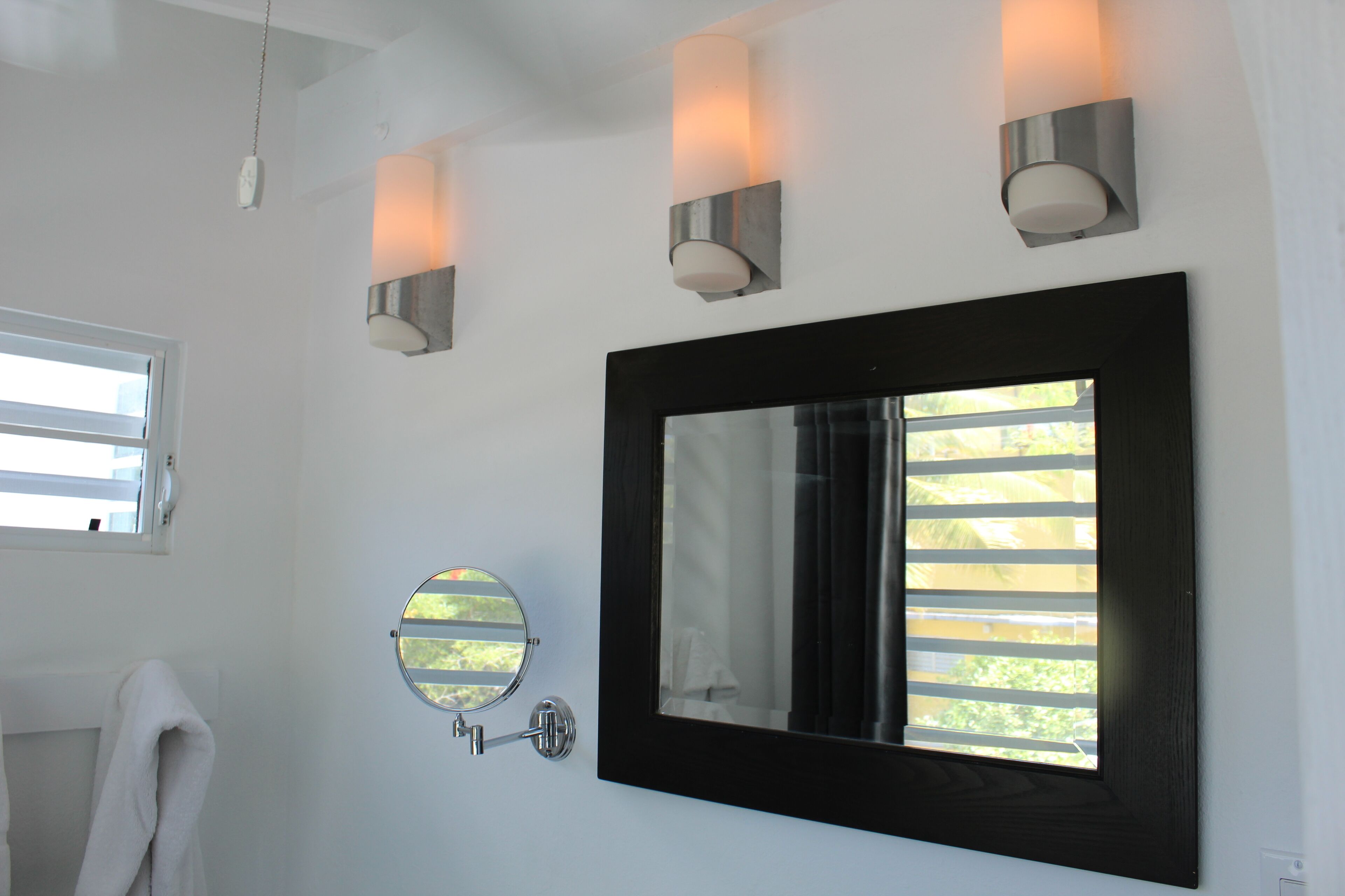 A bathroom with a large mirror, three wall-mounted lights, a round mirror, and a window with blinds.