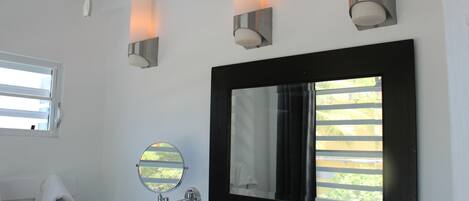 A bathroom with a large mirror, three wall-mounted lights, a round mirror, and a window with blinds.