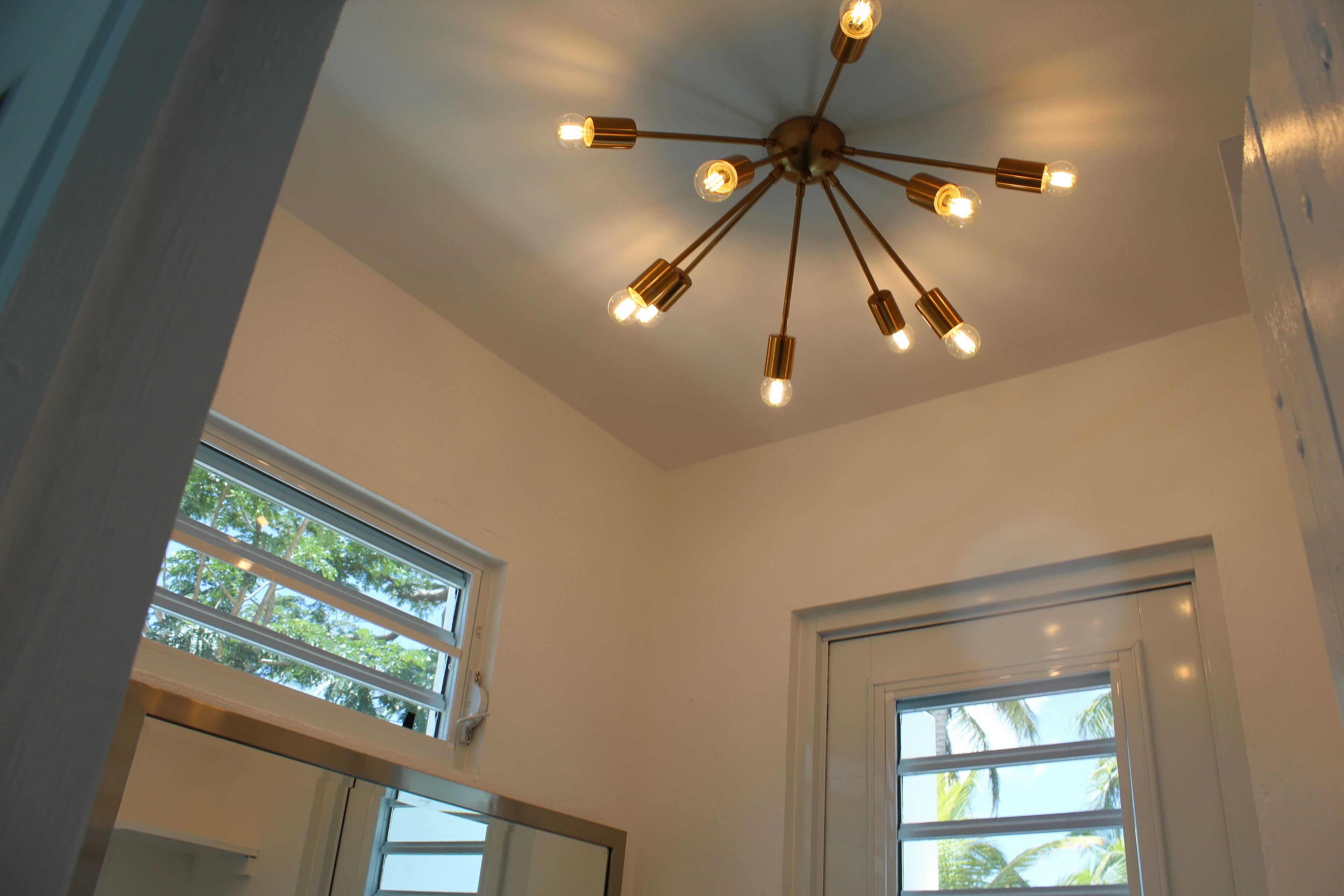 A room with a modern ceiling light fixture and two windows with views of palm trees.