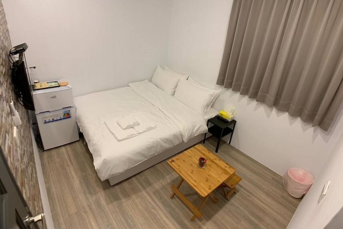 Une petite pièce, bien rangée, avec un lit, une table en bois, une table de chevet et un petit meuble.