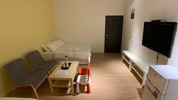 A compact hotel room with a bed, a sofa, a small table, a television, and a refrigerator.