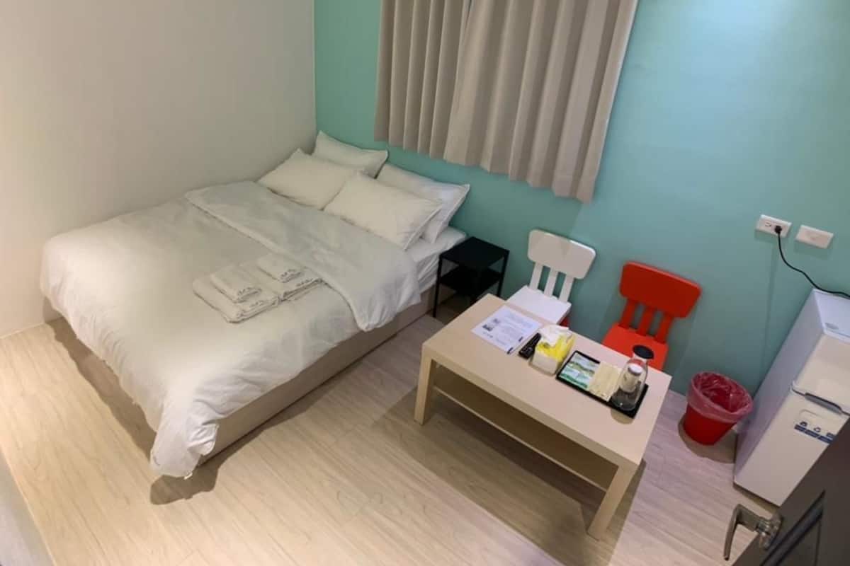 Une petite pièce, bien rangée, comprenant un lit, une table avec des articles de toilette, une chaise et un réfrigérateur.