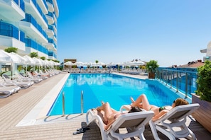 Un área junto a la piscina con sillones de descanso y sombrillas, un cielo azul despejado y un edificio con balcones.