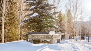 A snow-covered bench with a sign reading "Cottonwoods" in a park area with trees and a car in the background.