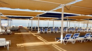 En strandnära paviljong med rader av vita solstolar under beige parasoller, ett trädäck och utsikt över havet.
