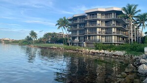 A multi-story hotel with balconies, located near a body of water with palm trees and a rocky shoreline.