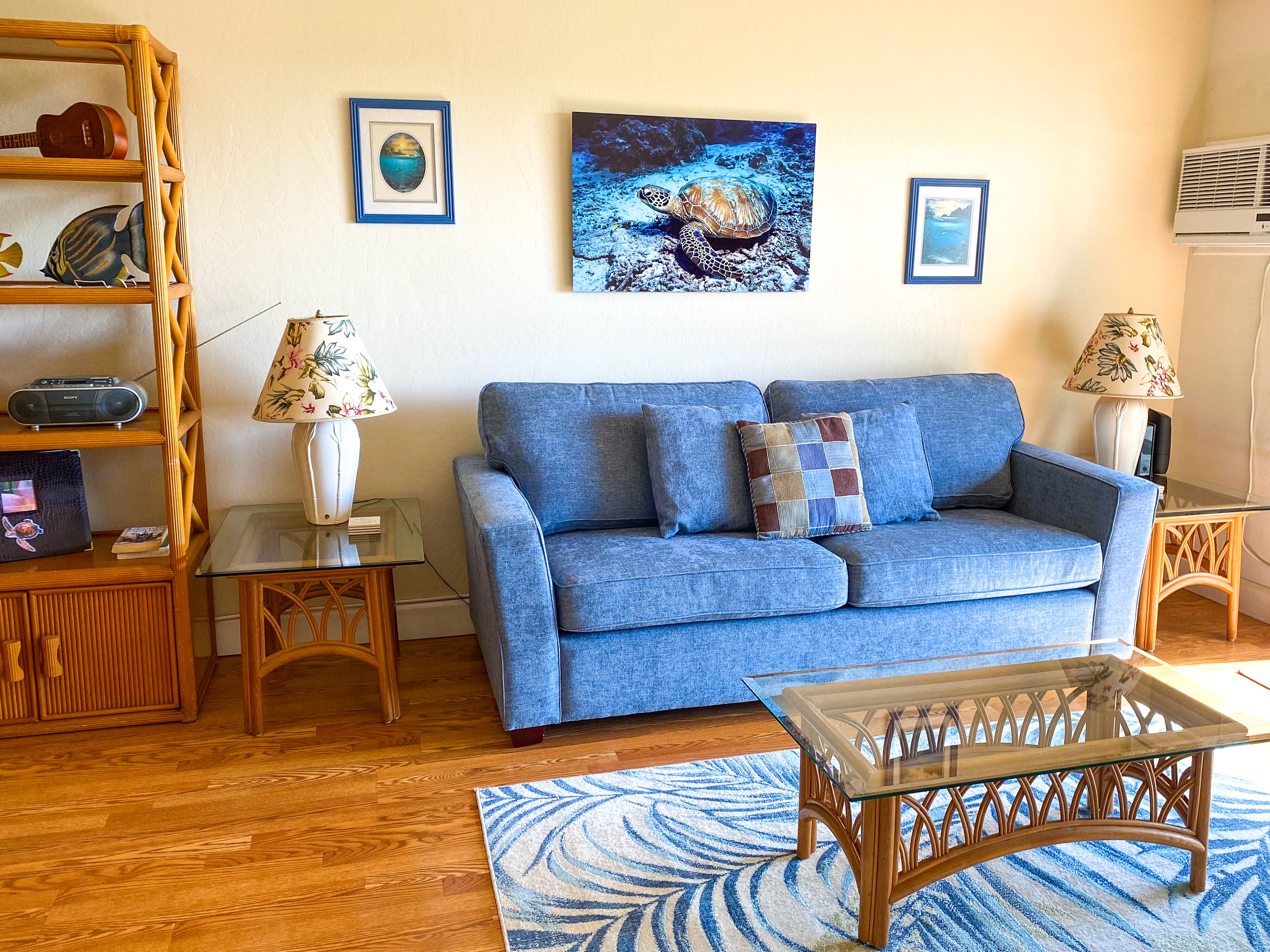 A living room with a blue sofa, two side tables with lamps, a glass coffee table, a wooden bookshelf, and framed pictures on the wall.