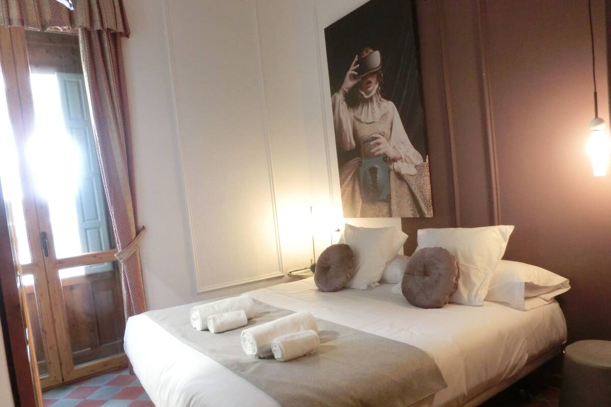 Een net opgemaakt bed met witte lakens en opgerolde handdoeken, een groot poster aan de muur, en een houten deur met gordijnen.
