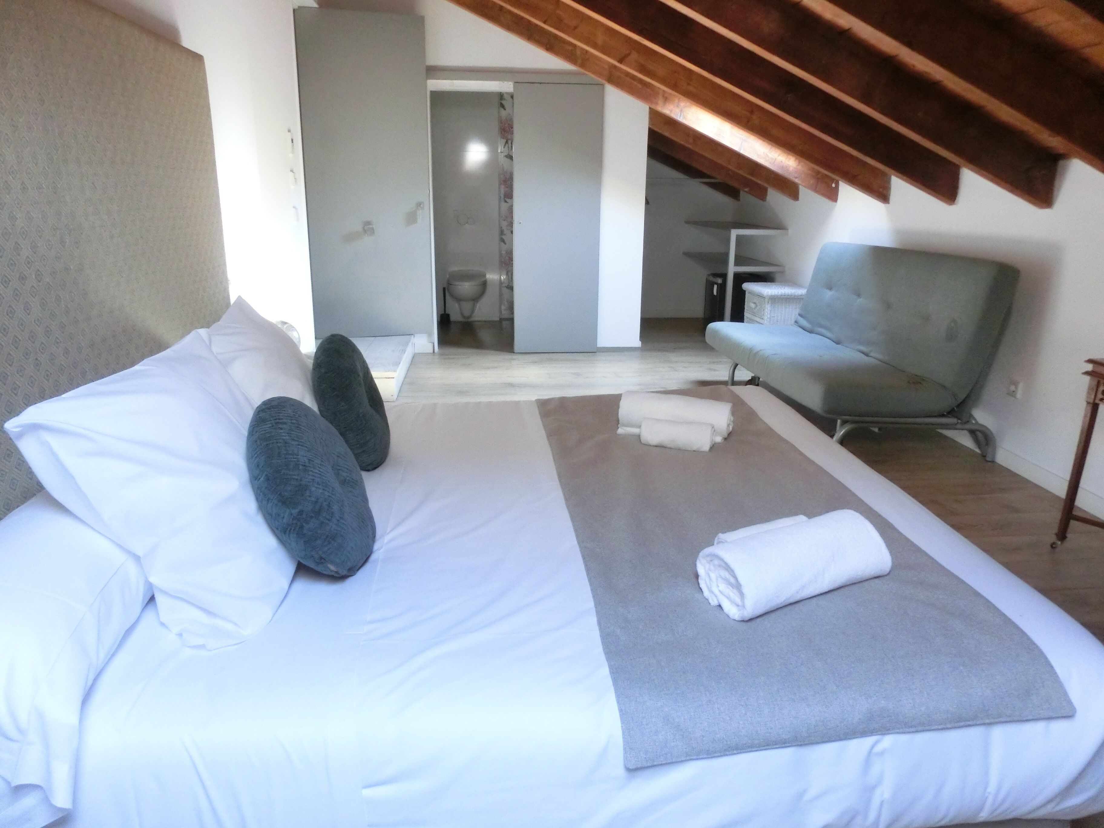 A bedroom with a large bed, a grey sofa, and a wooden ceiling.