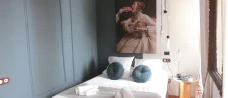 A bedroom with a bed, pillows, a painting, a bedside table, and a lamp.
