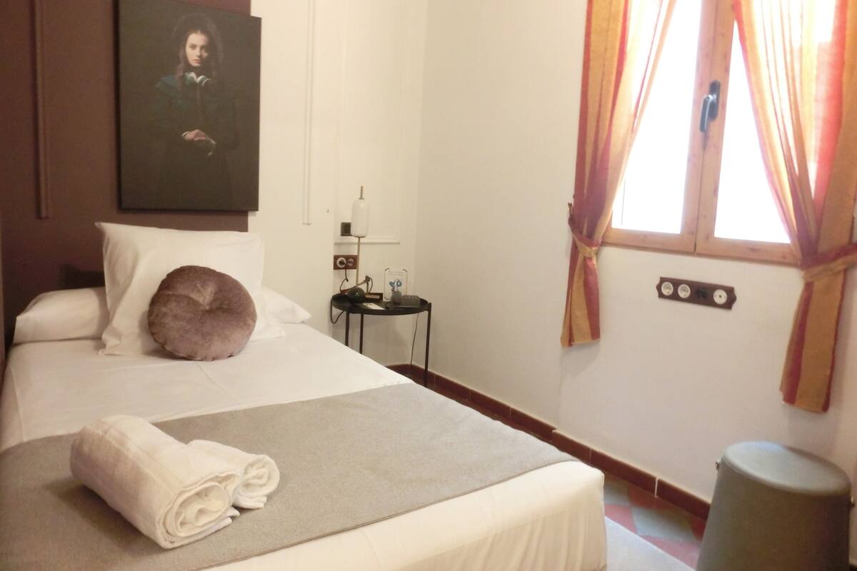 Een hotelkamer met een bed, een nachtkastje met een lamp, een schilderij aan de muur en een raam met gordijnen.