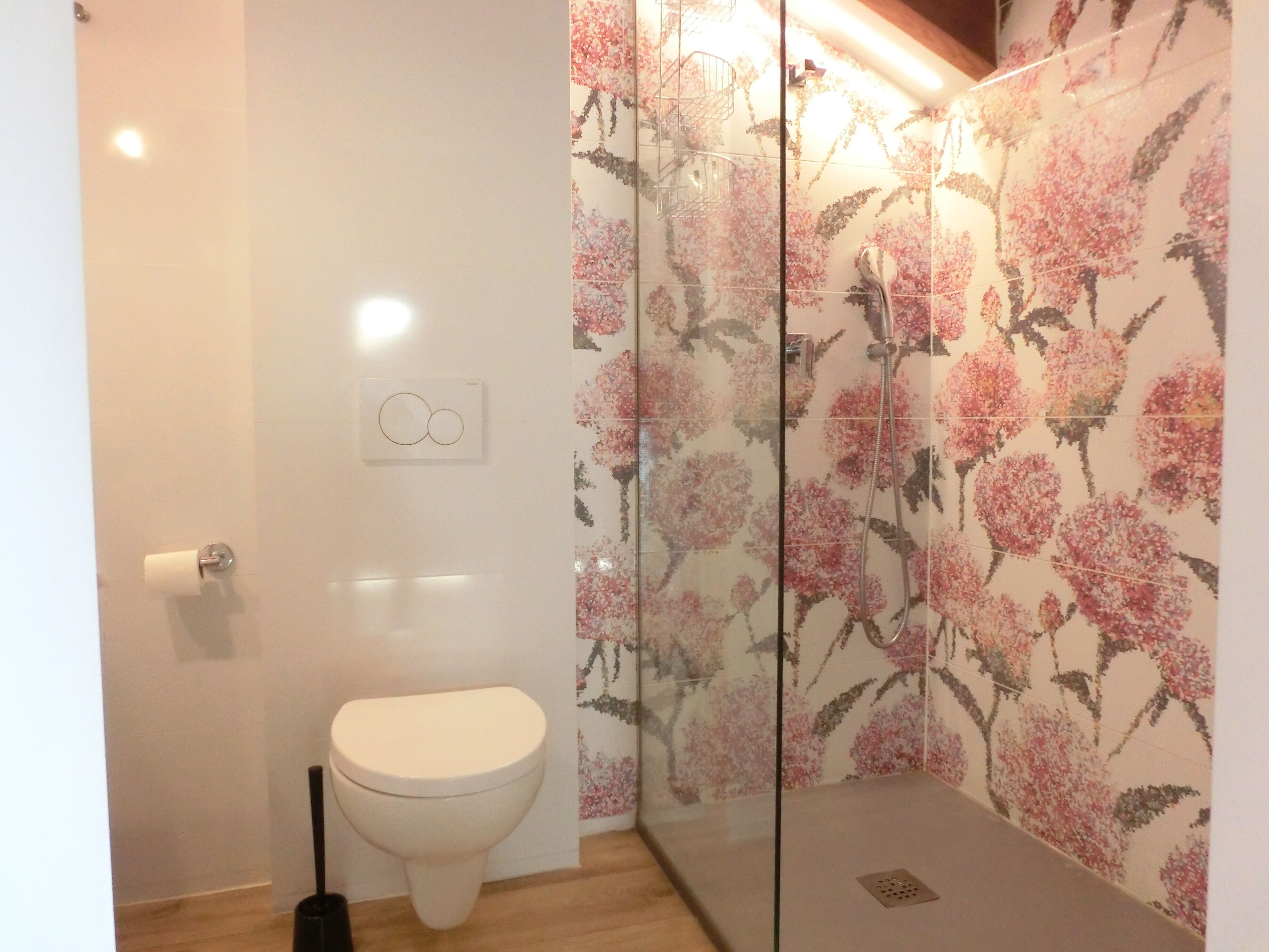 A bathroom with a toilet, a shower with floral tiles, and a glass partition.