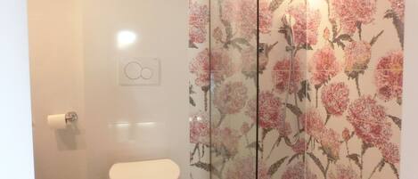 A bathroom with a toilet, a shower with floral tiles, and a glass partition.