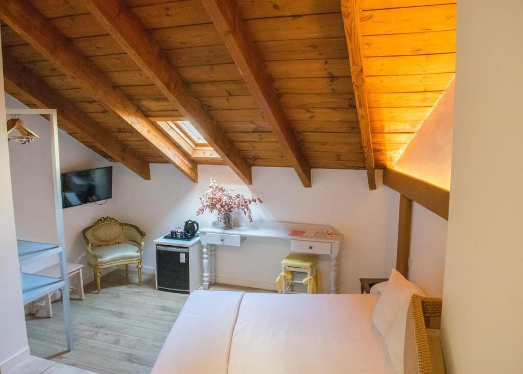 A cozy bedroom with a wooden ceiling, a bed, a desk with a chair, a TV, and a small table with a vase of flowers.