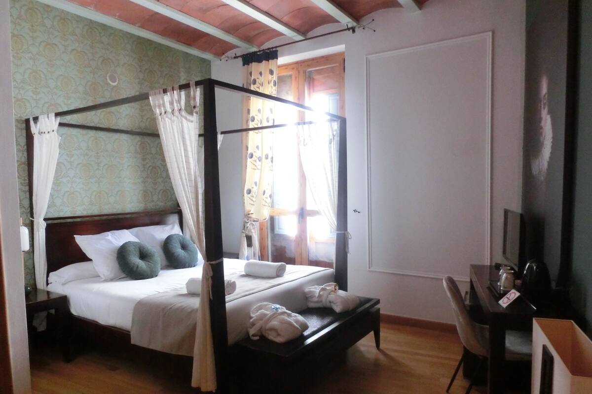 Een hemelbed met witte beddengoed en groene kussens, een houten bureau met stoel, en een televisie aan de rechterkant.
