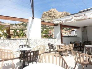 Een terras buiten met tafels en stoelen, een uitzicht op een heuvel met een gebouw, en een betegelde muur met een geometrisch patroon.
