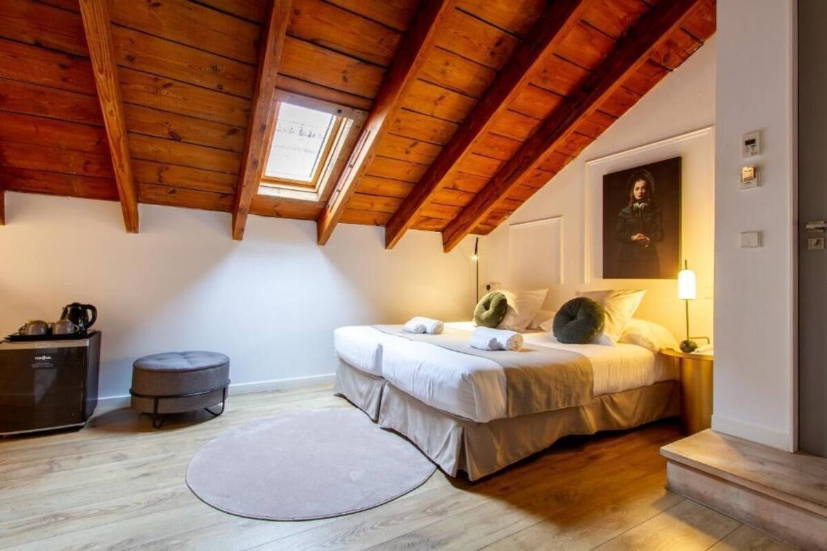 Een slaapkamer met een houten plafond, een bed met kussens, een nachtkastje met een lamp en een klein krukje.