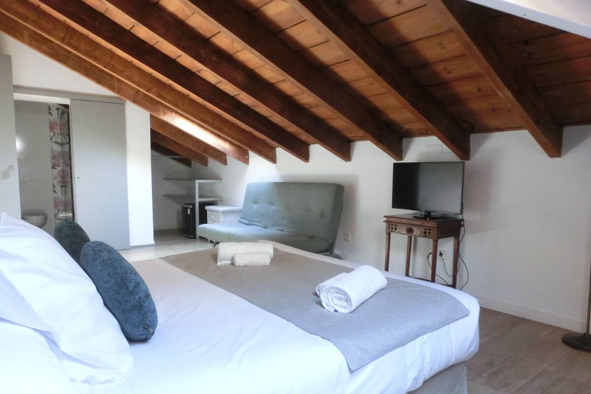 Een slaapkamer met een bed, een grijze bank, een televisie op een houten tafel en een houten plafond.