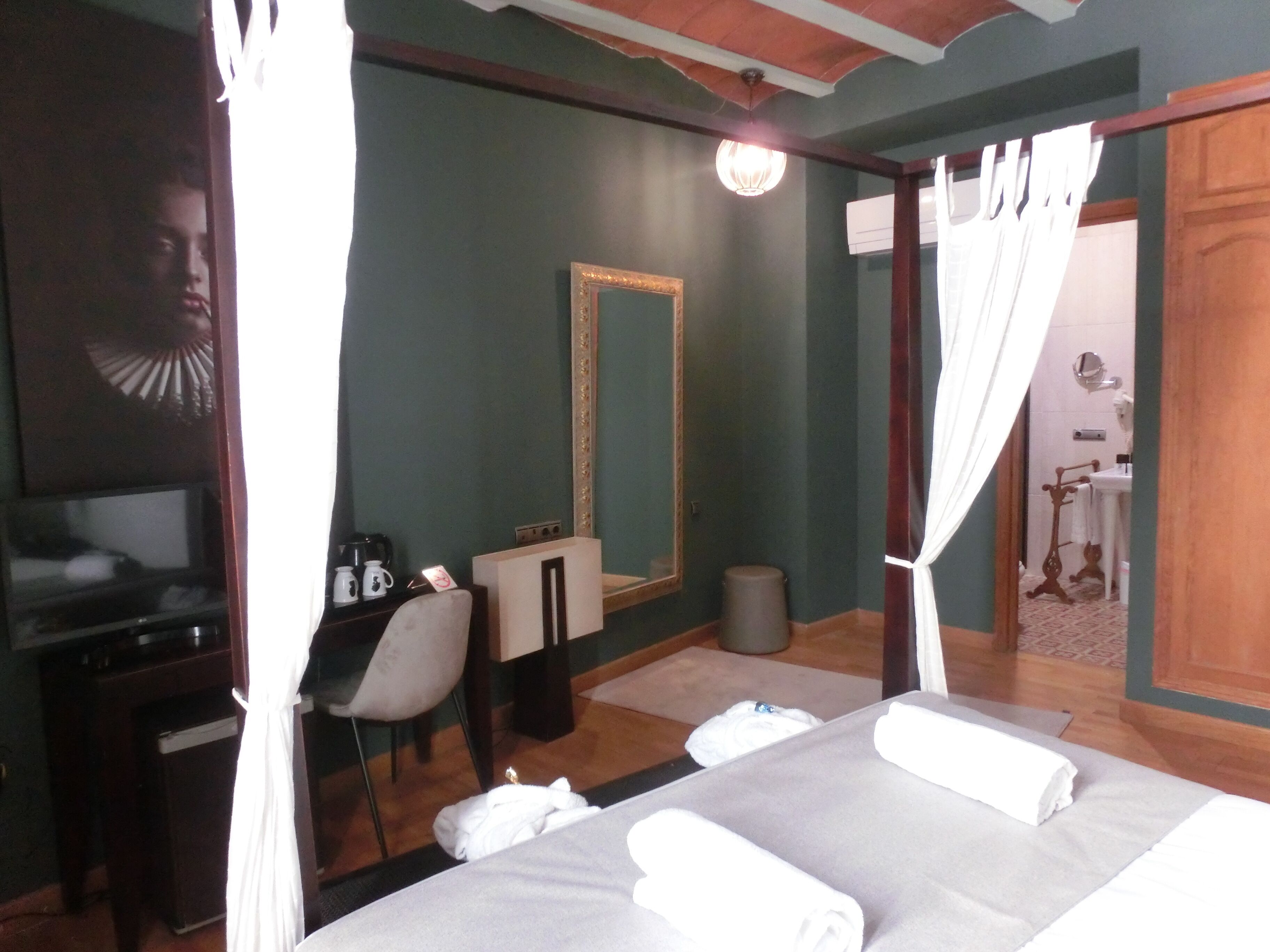 Habitación de hotel con cama, escritorio, silla, espejo y baño visible a través de una puerta abierta.