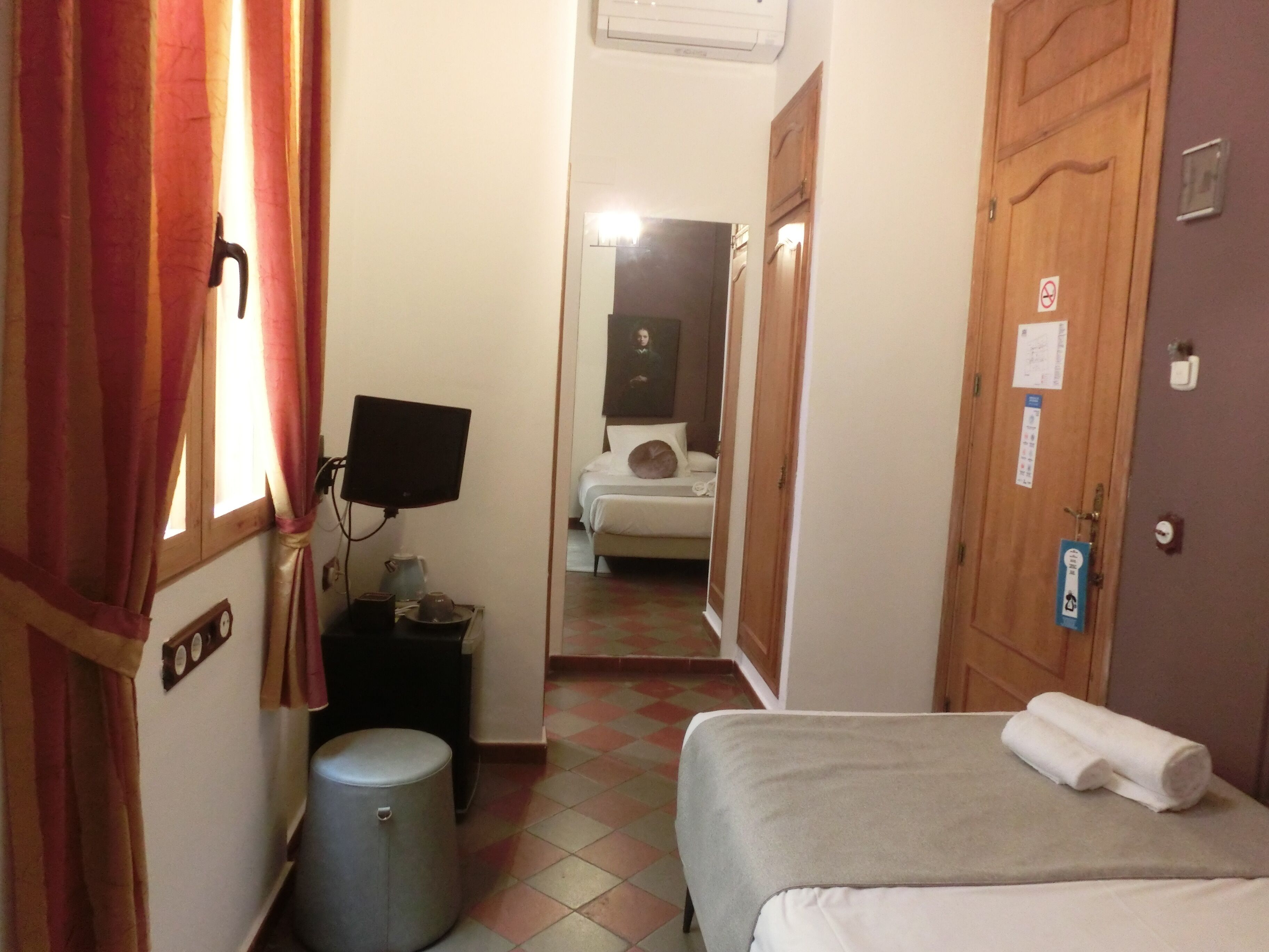 A hotel room with a bed, a small table with a kettle, a TV, a trash bin, and a bathroom visible through an open door.