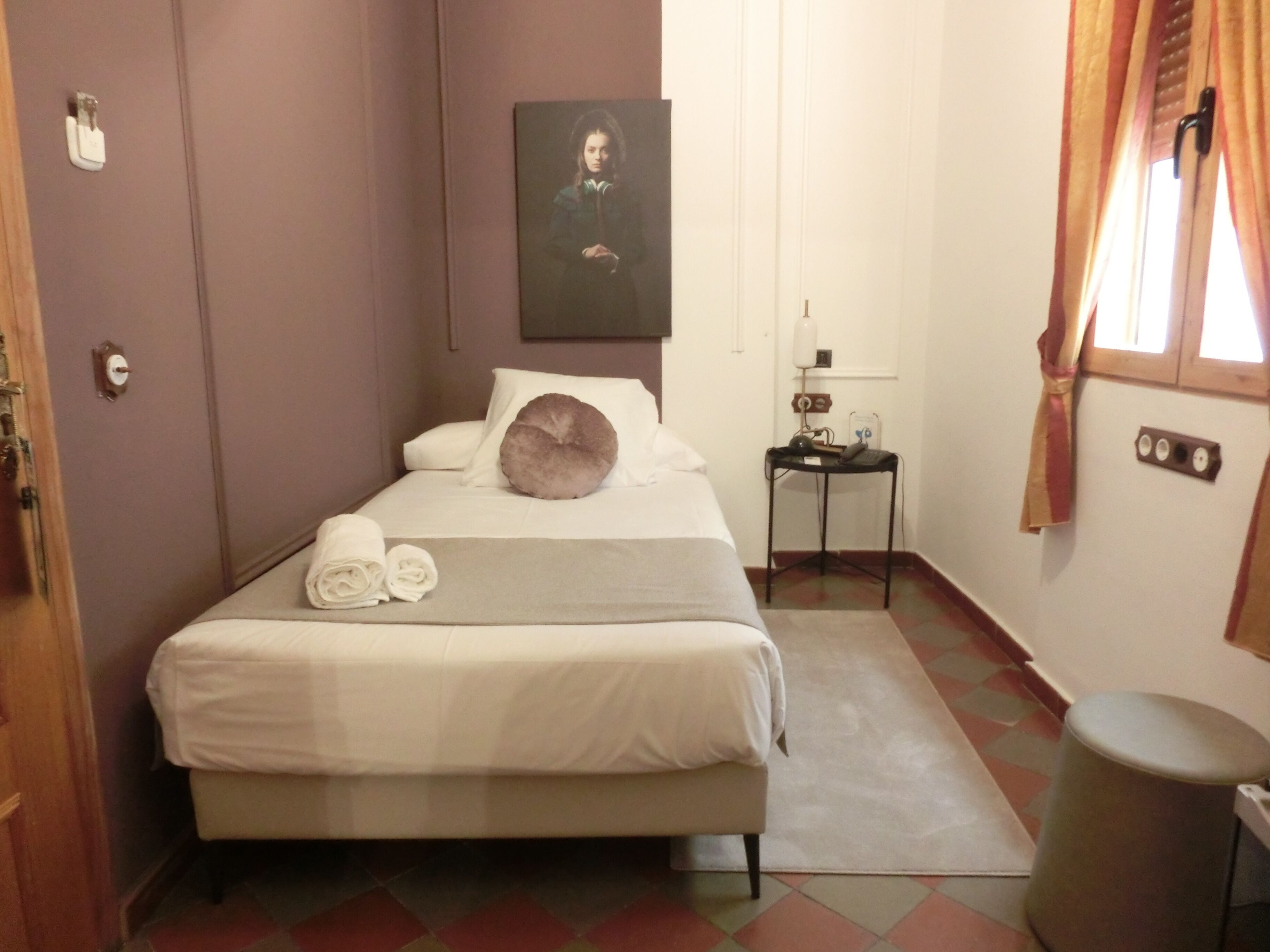 A neatly made bed with white linens and a gray pillow, a small bedside table with a lamp, and a trash can in a room with a checkered floor and a door on the left.