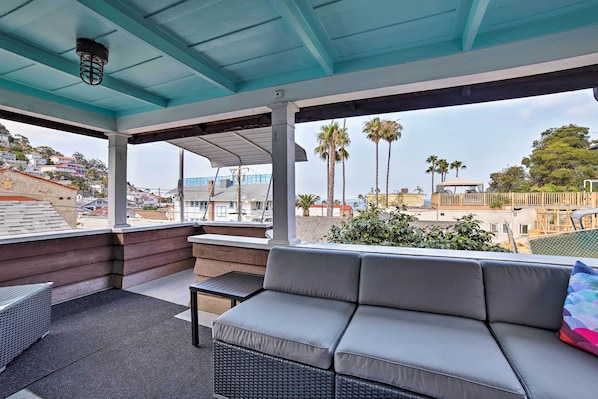 Un área de estar al aire libre techada con un sofá y una mesa, con vista a un barrio residencial con palmeras y casas.