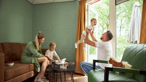 Une famille se trouve dans un salon, avec un canapé vert, une petite table ronde et une fenêtre avec des rideaux.