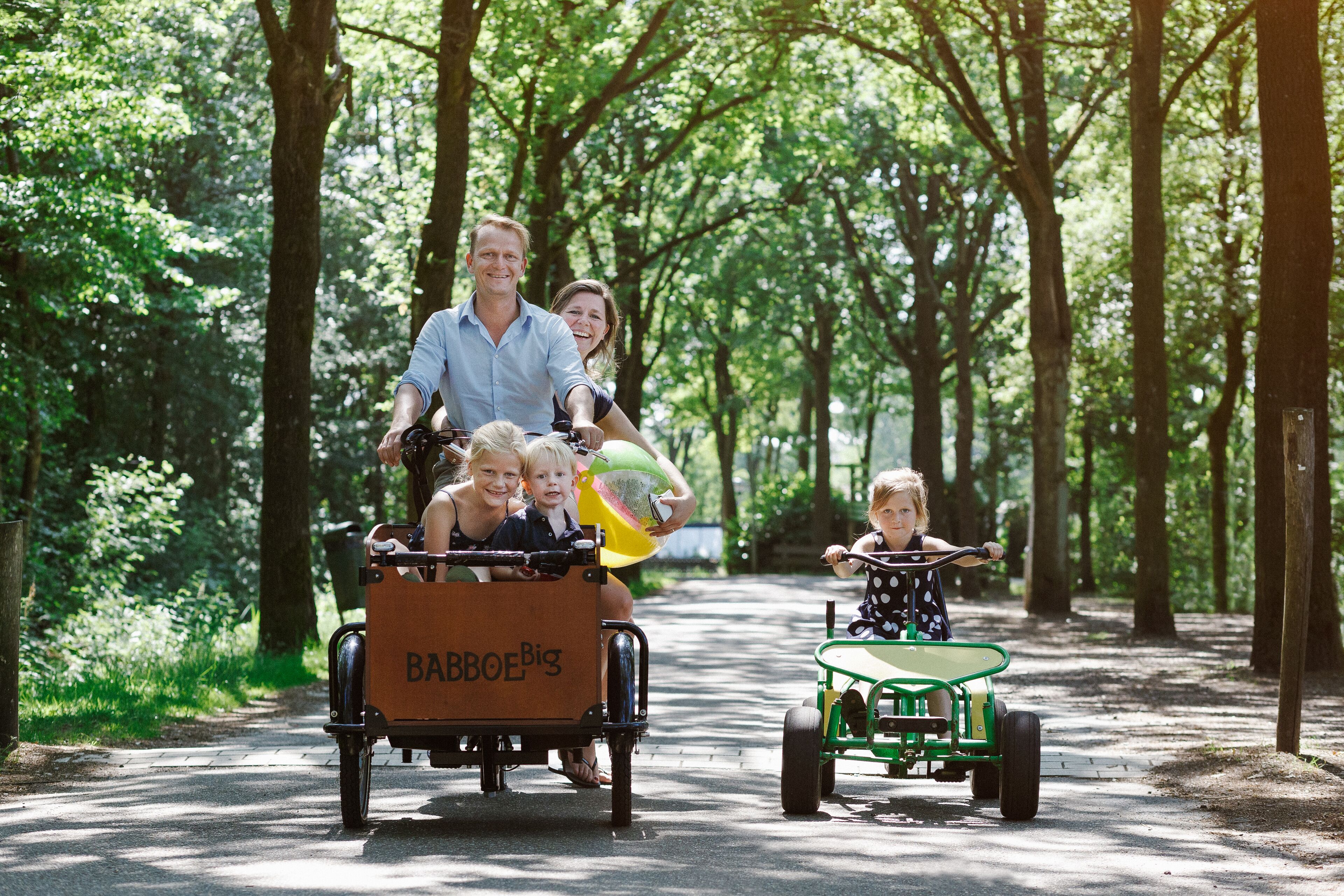 A family with a baby in a wagon and a child in a tricycle on a paved path in a wooded area.