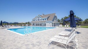 Une piscine avec des chaises longues et des parasols, une maison en arrière-plan et un ciel dégagé.