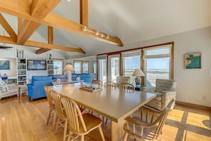 Un vaste espace de vie comprenant une grande table à manger en bois, un canapé bleu et des chaises en bois. De nombreuses fenêtres laissent entrer la lumière naturelle et offrent une vue sur l’extérieur.