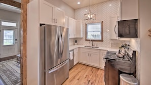 A modern kitchen with white cabinets, a stainless steel refrigerator, a herringbone-patterned backsplash, a hanging light fixture, and a window with blinds. - Updated Indianapolis Home, Walk to Lucas Oil! (Indianapolis)