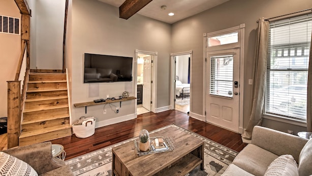 A spacious living room with a wooden staircase, a flat-screen TV mounted on the wall, a coffee table with a decorative item, and a large window with blinds. - Updated Indianapolis Home, Walk to Lucas Oil! (Indianapolis)
