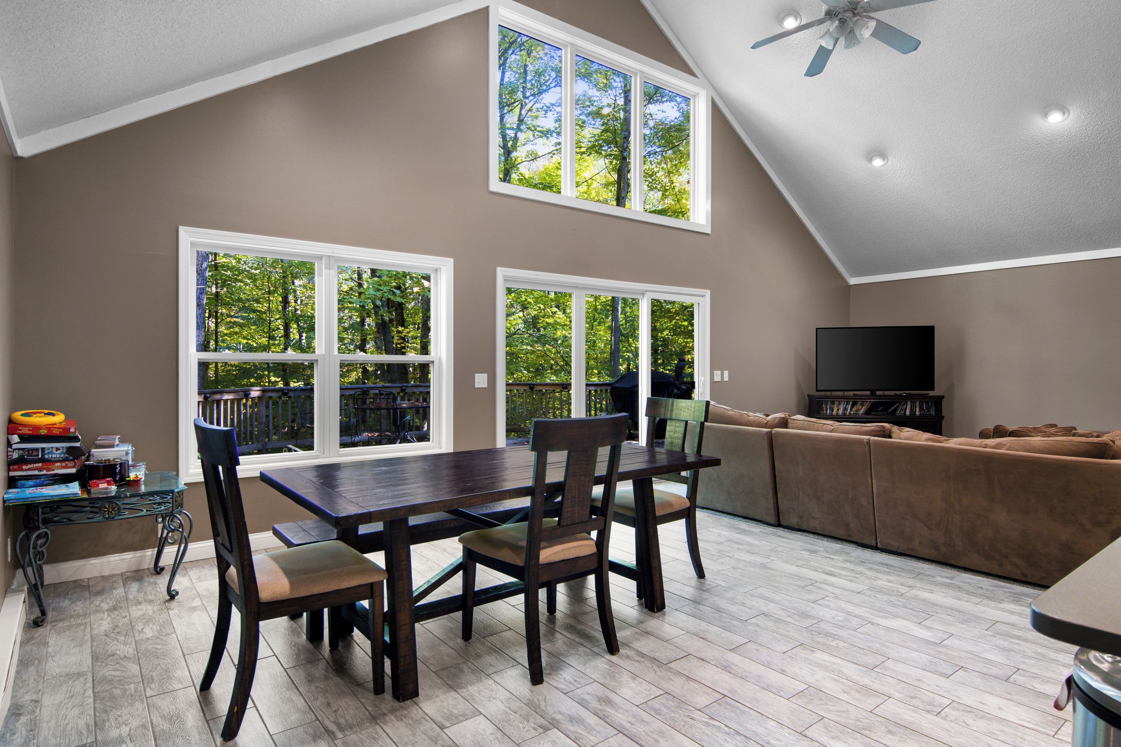 A dining area with a table and chairs, a sofa, and a TV in a room with large windows and a ceiling fan.