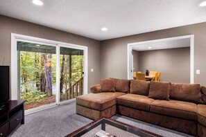 A living room with a brown sectional sofa, a glass coffee table, and a view of a forest through a sliding glass door. - Schuss Mountain Chalet (Mancelona)