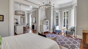 A spacious bedroom with a large bed, a sitting area with a chair and a table, a fireplace, and a view of the outdoors through large windows. - Mayor's luxury 1869 historic home for 4 on the square! (Savannah)