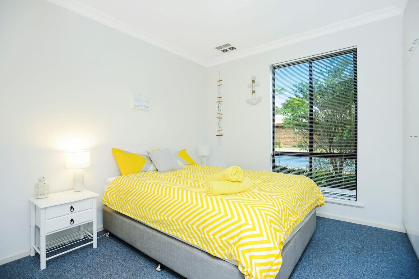 Un dormitorio con cama, mesita de noche, lámpara, ventana con persianas y vista a zonas verdes.