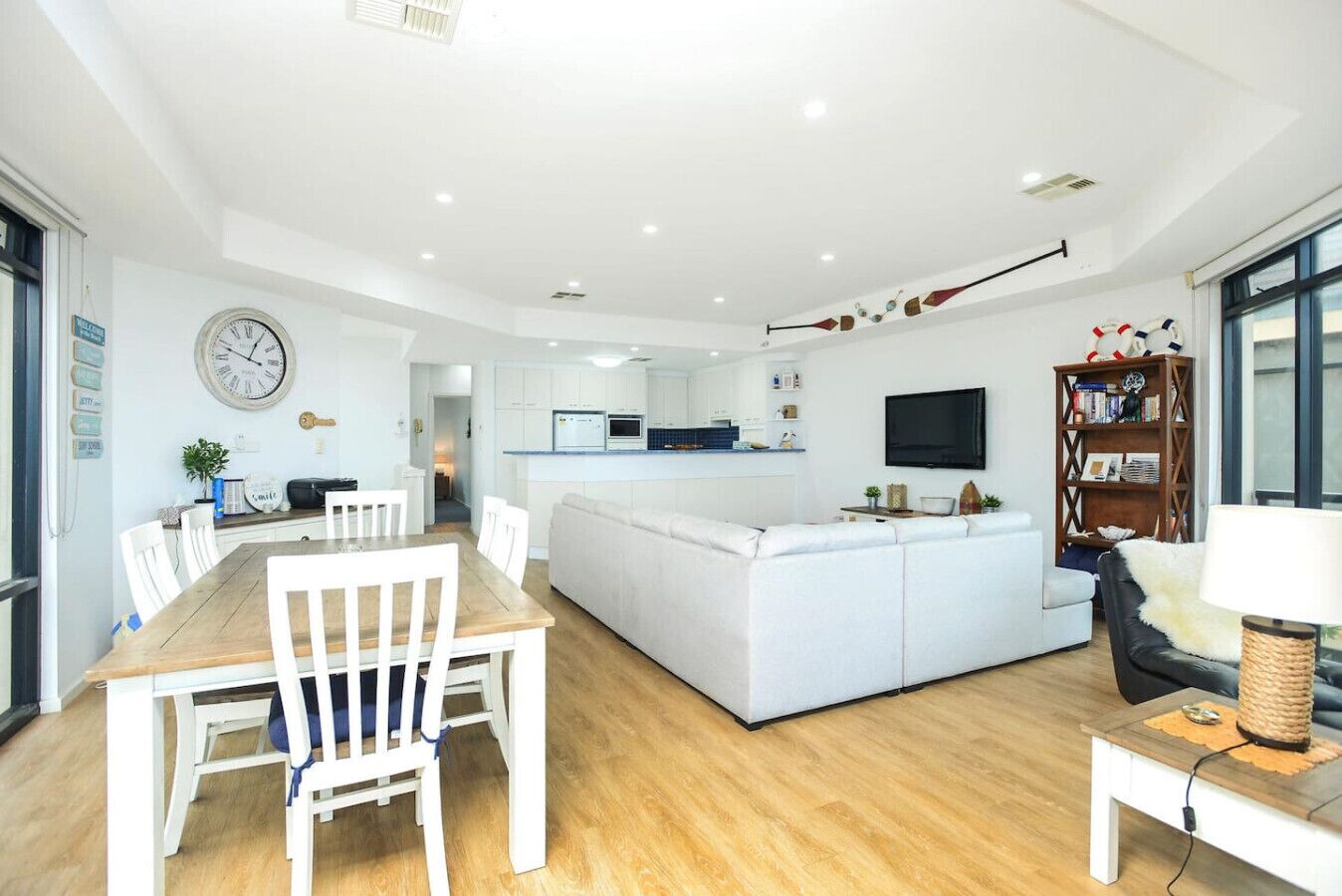 A modern living room with a white sofa, a dining table with chairs, a kitchen area, and a bookshelf.