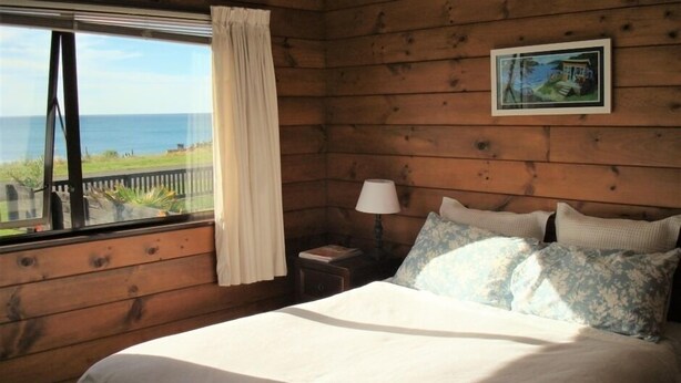 A wooden cabin room with a bed, a nightstand, a lamp, a window with a view of the ocean, and a framed picture on the wall.
