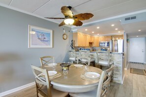 Un coin repas avec une table ronde dressée pour quatre personnes, entourée de chaises en bois. Un ventilateur de plafond orné de motifs de feuilles est visible au-dessus de la table.