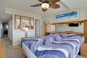 Une chambre à coucher comprenant un lit, un ventilateur de plafond, une table de chevet, une lampe, un téléviseur et un tableau représentant une scène de plage.