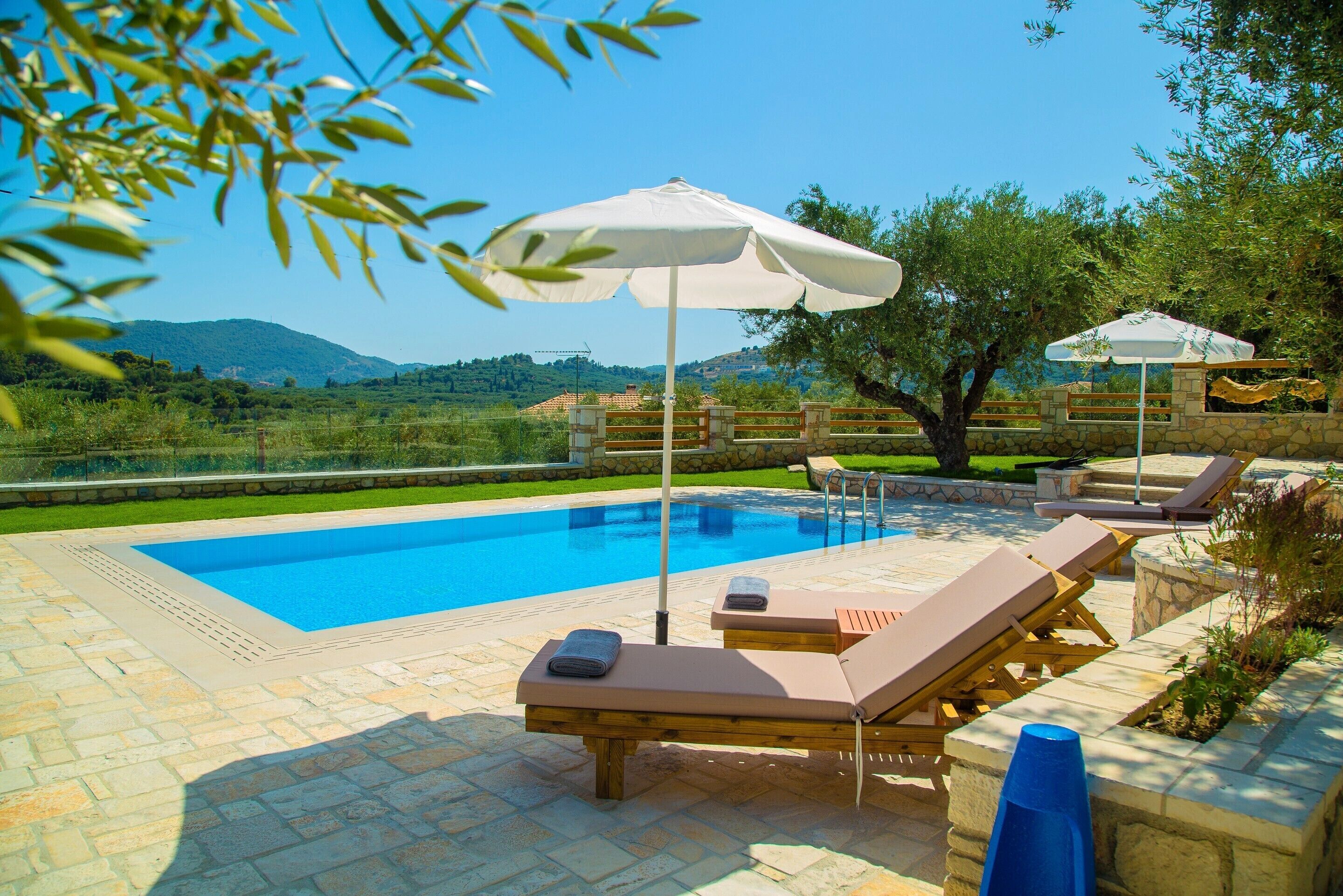 A swimming pool with lounge chairs and umbrellas, surrounded by stone tiles and greenery.