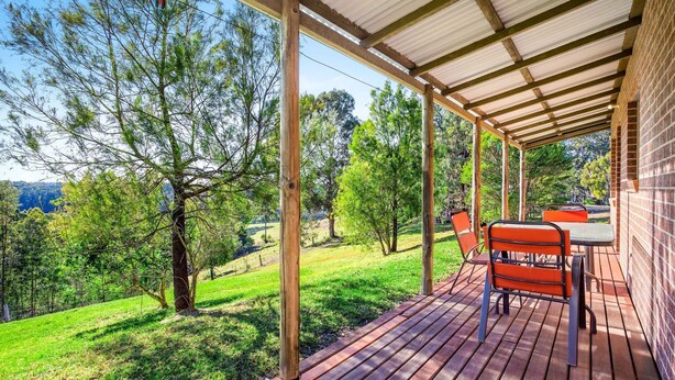 A covered outdoor area with a wooden deck, a table, and chairs, surrounded by trees and a view of the landscape. - Braeside Cabin Two - Alphitonia (CENTRAL TILBA)