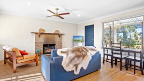 A living room with a fireplace, a sofa, a wooden chair, a dining table with chairs, and a large window with a view of trees. - Braeside Cabin Two - Alphitonia (CENTRAL TILBA)